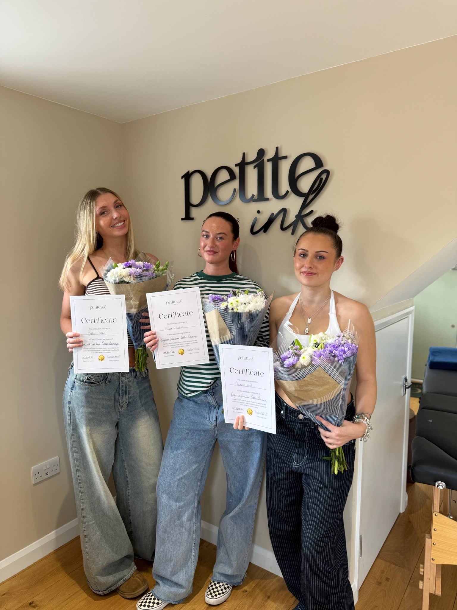 Three graduates with certificates and flowers under the Petite Ink sign